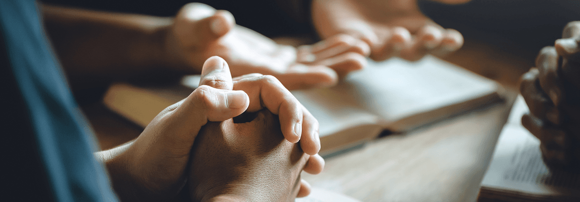 Close-up of hands clasped in prayer with open books on a table.