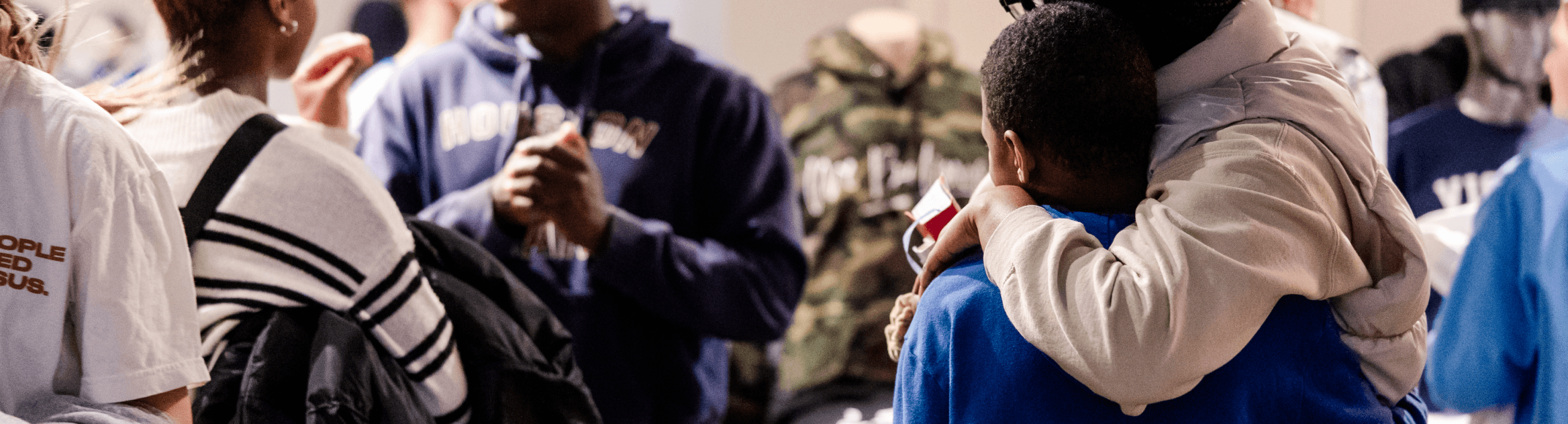 A group of people stand close together in a crowded indoor setting. One person has their arm around a child, while others talk nearby.